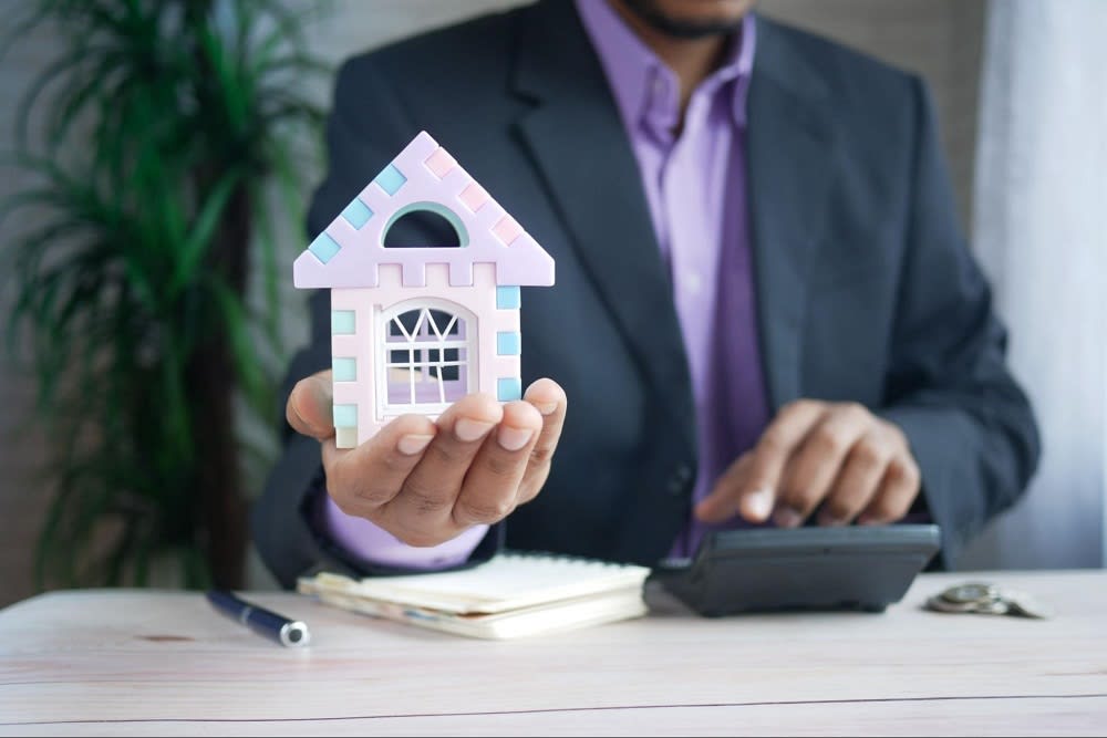 A person in a suit holds a small pastel-colored house model while using a calculator on a desk.