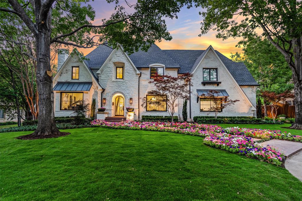 A Colonial-style white house with a black slate roof, chimney, covered porch, walkway, front lawn, trees and flowers.