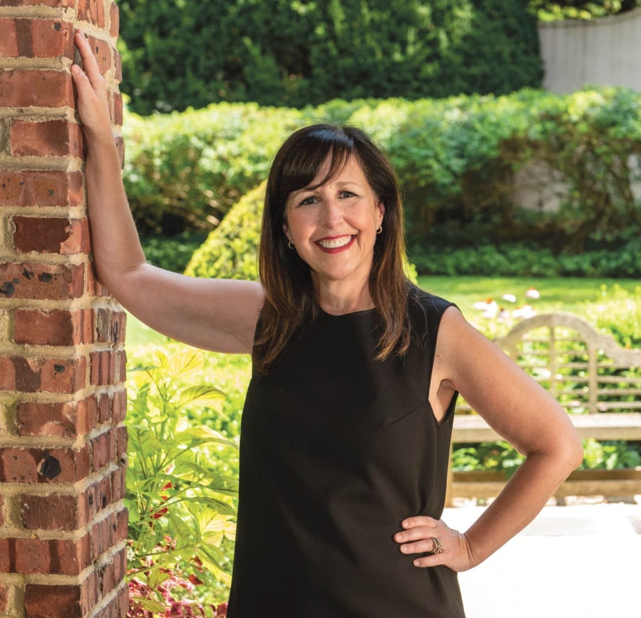 A woman with brown hair wearing a black sleeveless top leans against a brick wall, wearing earrings.