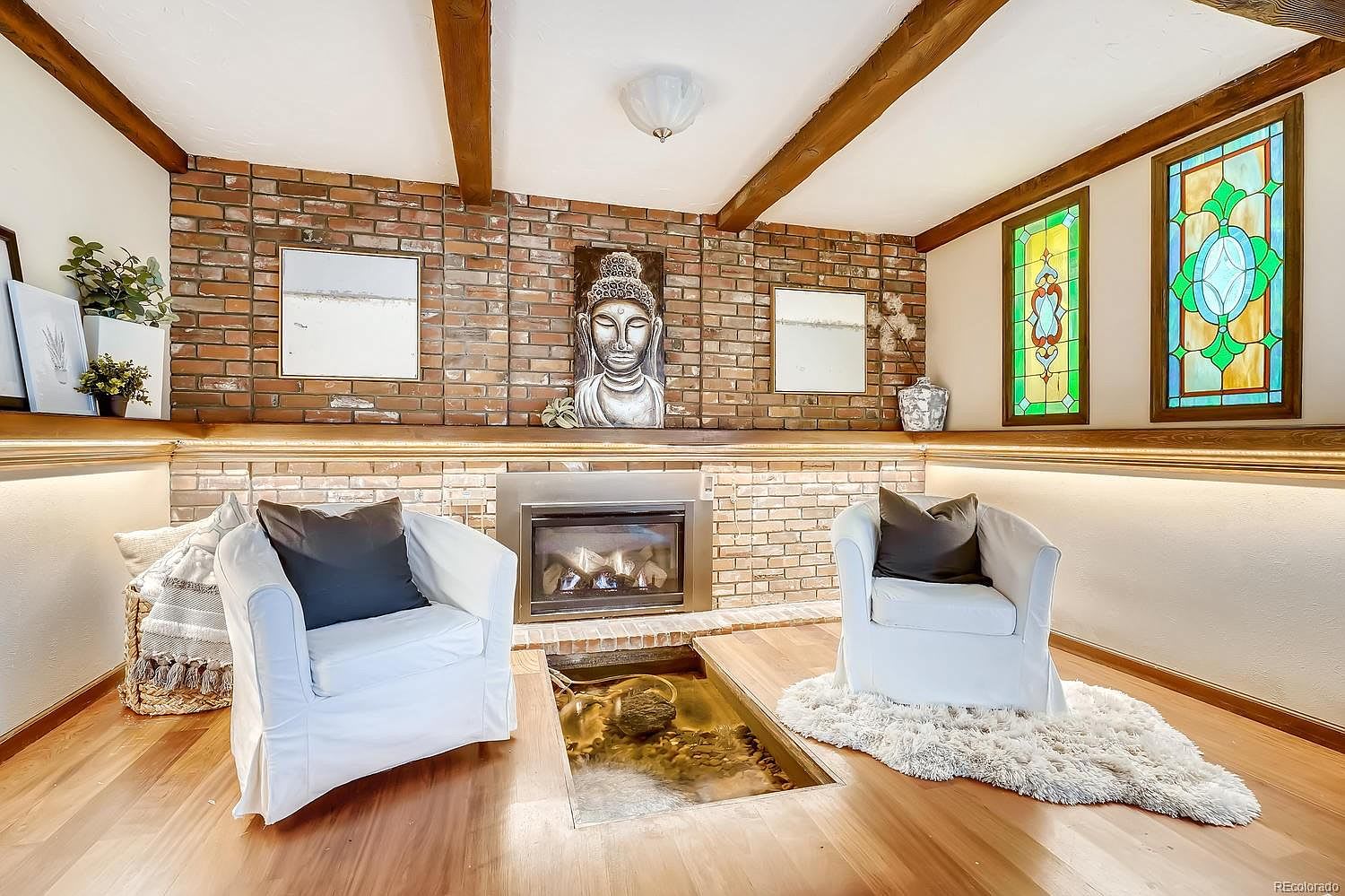 Living room with a brick fireplace, stained-glass windows, exposed beams, and two white armchairs.