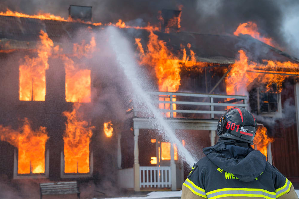 House engulfed in flames as a firefighter sprays it with water.