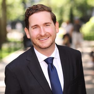 Jeffrey Kelly in a black suit and navy tie against a softly blurred outdoor background.