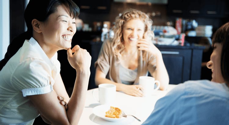 Three women enjoy coffee and cake together at a table, sharing laughter and conversation in a cozy setting.