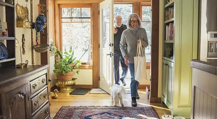 An elderly couple with a dog standing walking in an a well-decorated entryway or hallway. The space has a warm, inviting feel with various decorative items and plants.