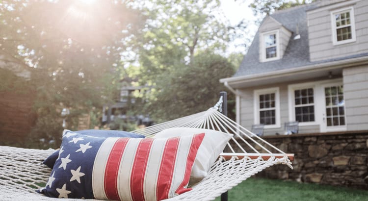 American flag pillow on a hammock sitting in the front yard of a traditional suburban home with dormer windows