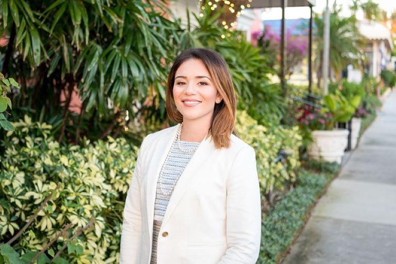 Woman in a white blazer standing on a sidewalk with greenery in the background
