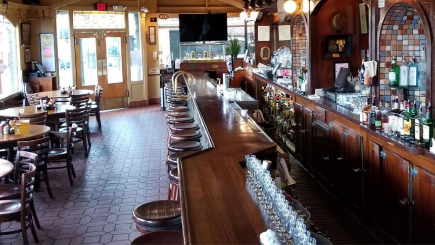 Interior view of the Buena Vista Cafe in Russian Hill, featuring round wooden tables with chairs, a long bar counter with stools, and a variety of liquor bottles displayed against a mirrored backdrop. The cafe exudes an old-fashioned charm with its hexagonal tiled floor, warm lighting, and walls adorned with mirrors and framed artwork.