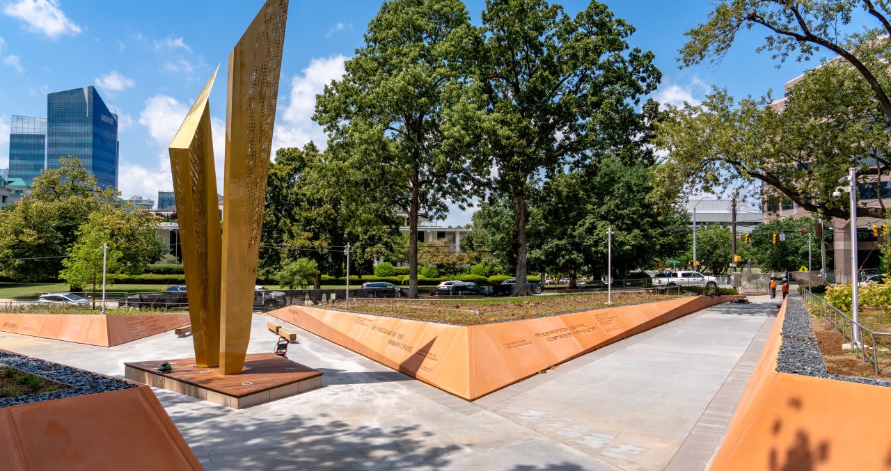 A modern outdoor memorial featuring a tall golden sculpture, surrounded by landscaped areas and pathways.