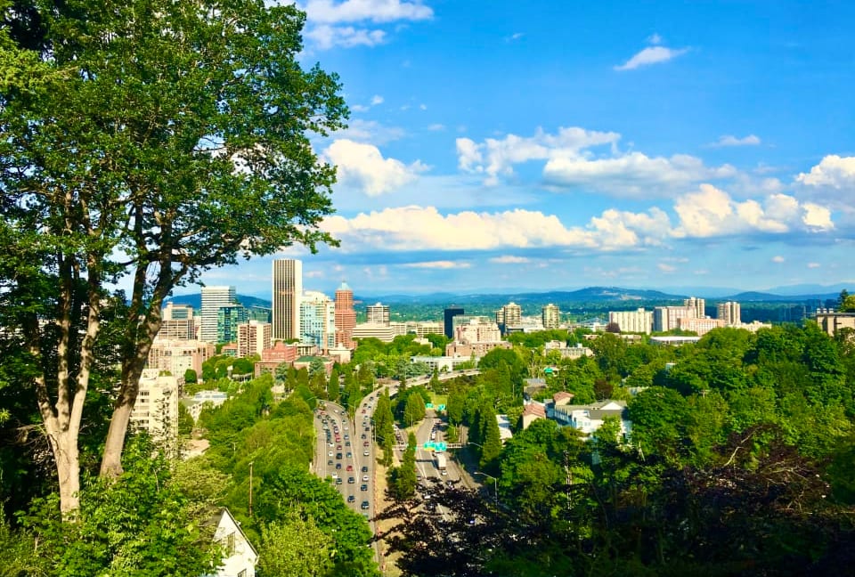 A view of the city in Portland from a hilltop. The city is surrounded by trees and mountains.