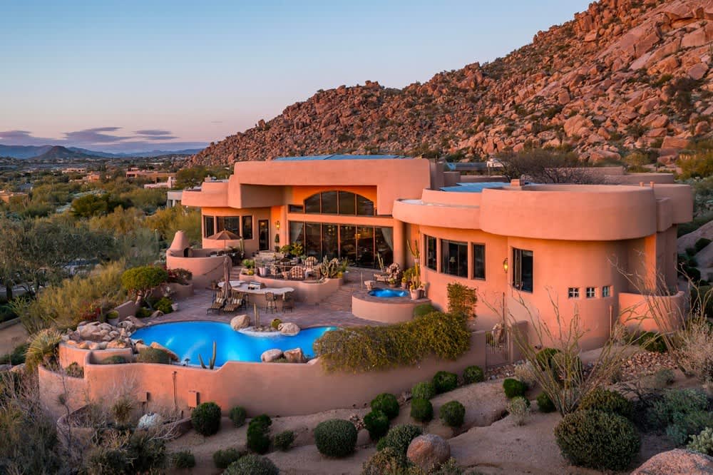 Luxury desert home with curved architecture, large windows, and a swimming pool nestled against a rocky hillside.
