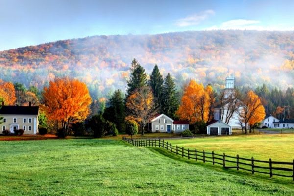 A green field with homes and a church in the background in the village of Tyringham, Massachusetts. The trees and a mountainous backdrop are dressed in brilliant fall colors.