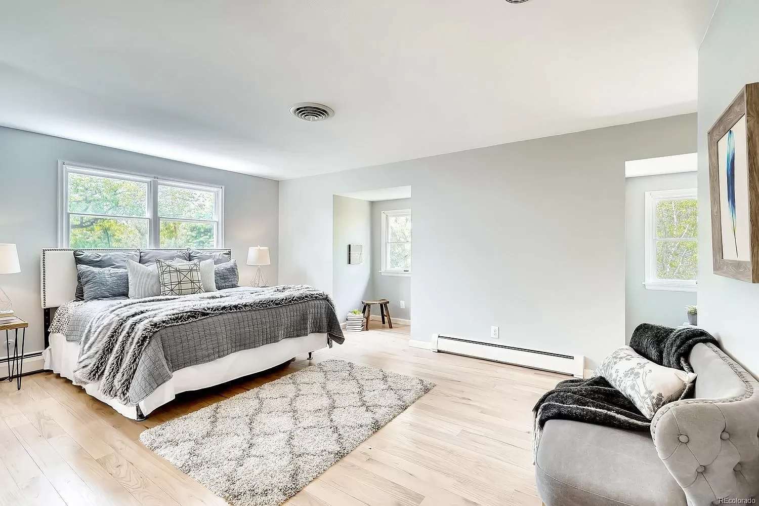 Bright bedroom with a large gray-and-white bed, pale wood floors, and a gray sofa.