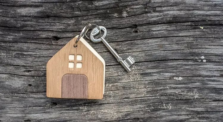 A small wooden house keychain with a metal key attached, set on a wooden surface. This image symbolizes home ownership or real estate.