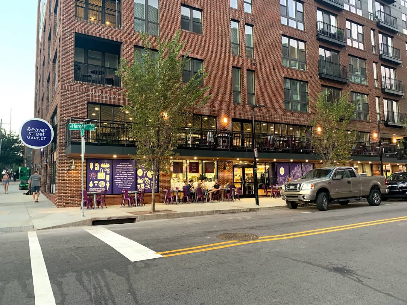 A brick building with a market sign, outdoor seating, and trees. People walk by, and cars are parked on the street.