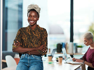 Smiling woman stands with arms crossed in a bright office as another woman works at a table.