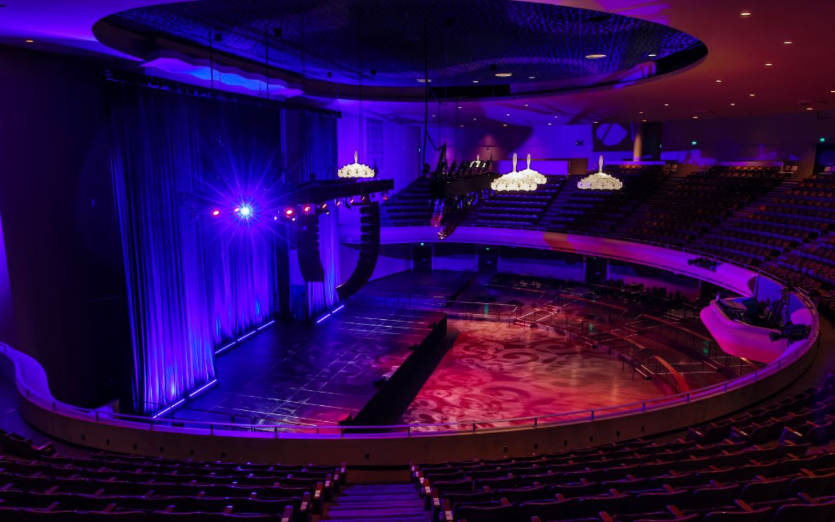 The grandeur of the Masonic Auditorium unfolds within its spacious interior. Elegant chandeliers cast a soft glow, illuminating the royal blue curtains framing the large stage. Multiple levels of empty seating areas await an audience, while intricate ceiling designs add to the venue’s aesthetic appeal.”