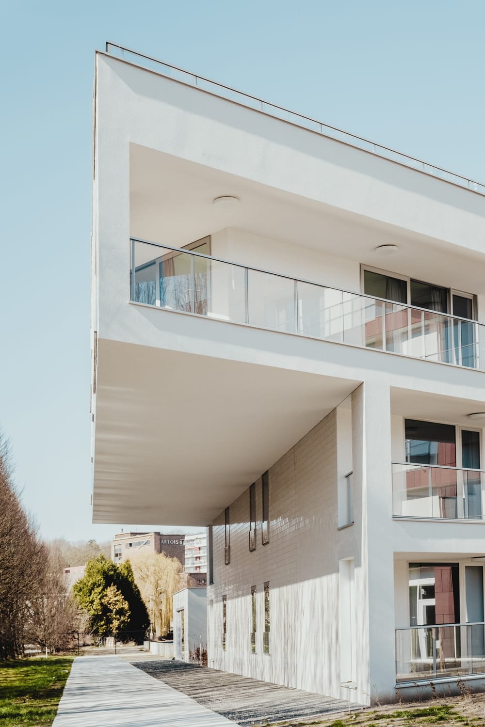Modern white apartment building with glass balconies and a long overhanging roof beside a walkway.