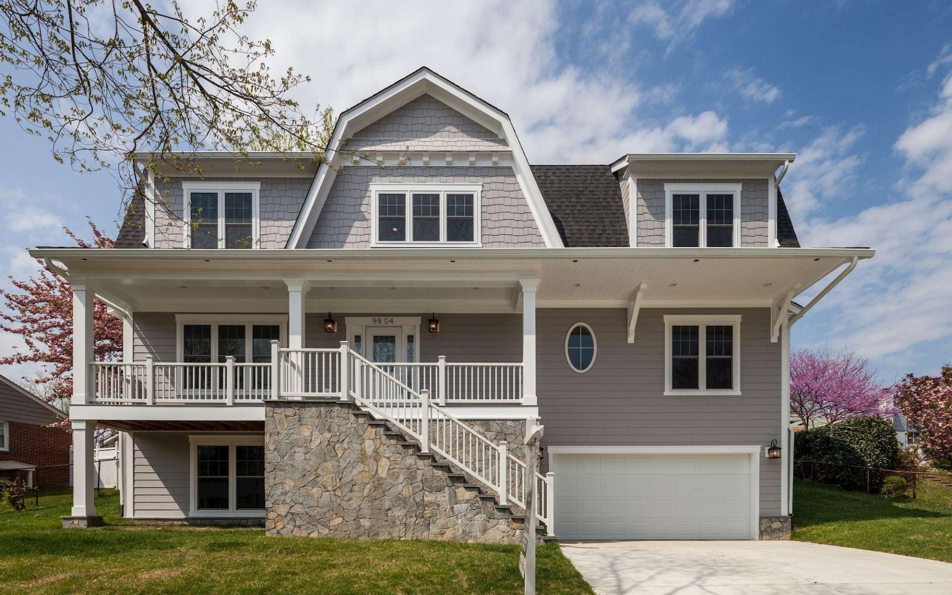 Gray house with white trim, a covered porch, stone staircase, dormers, and an attached garage.
