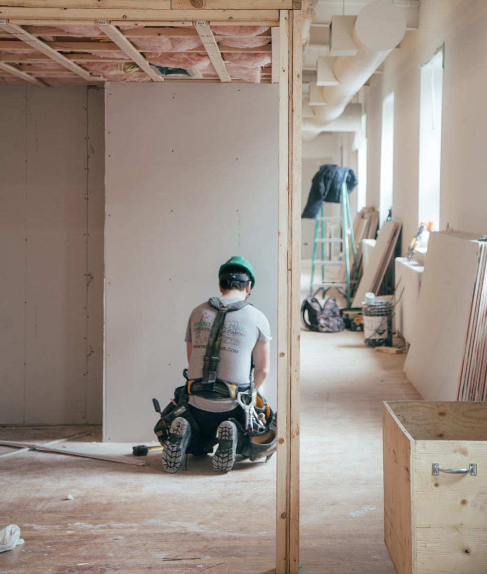 Unfinished home interior with exposed insulation and a worker kneeling beside drywall.