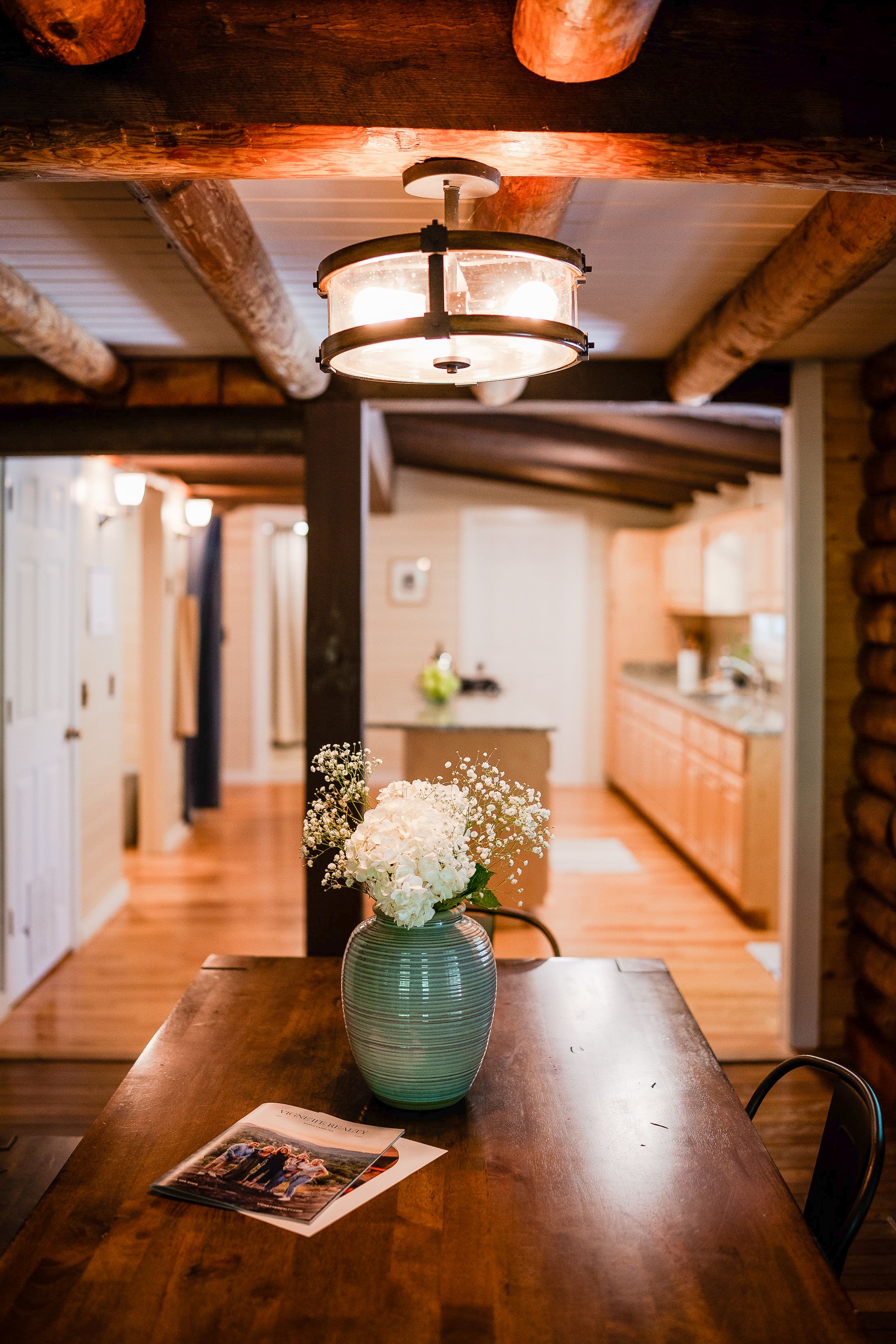 A cozy dining area with a wooden table, a vase of white flowers, and a magazine. Overhead, a rustic light fixture illuminates the space, showcasing the inviting atmosphere that real estate agents often highlight. The background reveals a warmly designed wooden kitchen.