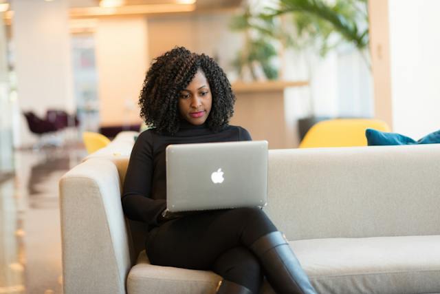 A woman dressed in black sitting on a couch with a laptop in her lap.