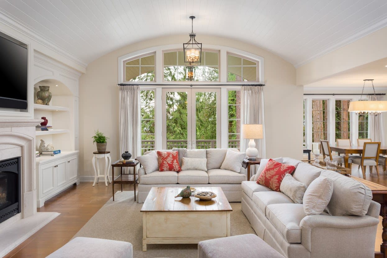 Living room with various furniture with cream-color décor, a stone fireplace, a dark wooden mantelpiece, and large windows.