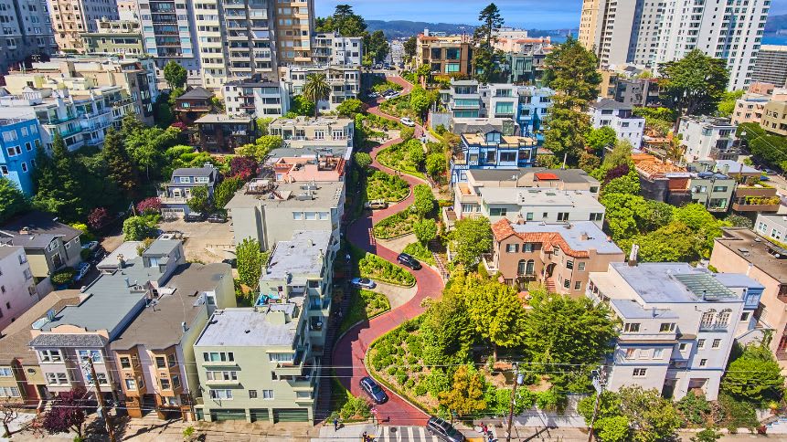 Aerial view of Lombard Street in San Francisco, showing its iconic switchbacks and red brick pavement. The street winds through a lush green hillside with trees and flowers in bloom. Cars navigate the curves, and pedestrians walk on the sidewalk.