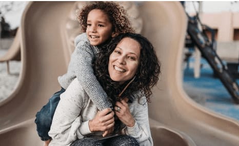A woman carries a smiling child on her back beside a curved play structure.