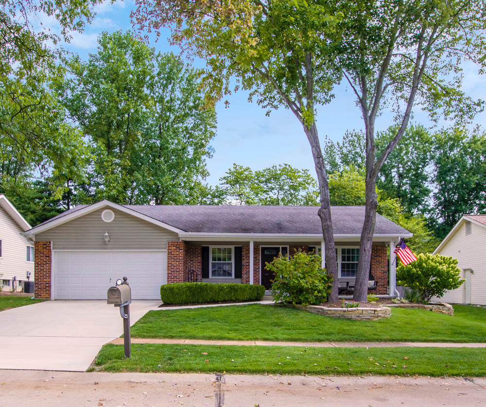 Single-story home with a two-car garage, brick accents, mature shade trees, and an American flag by the porch.