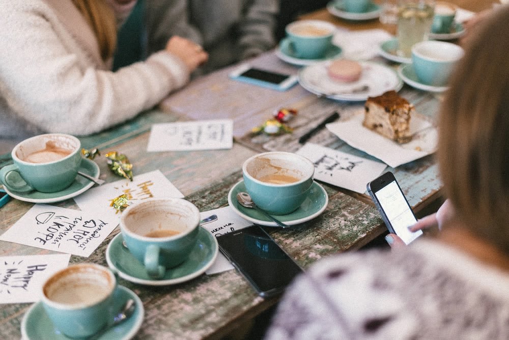 People share coffee around a worn café table with turquoise cups, cake, phones, and handwritten cards.