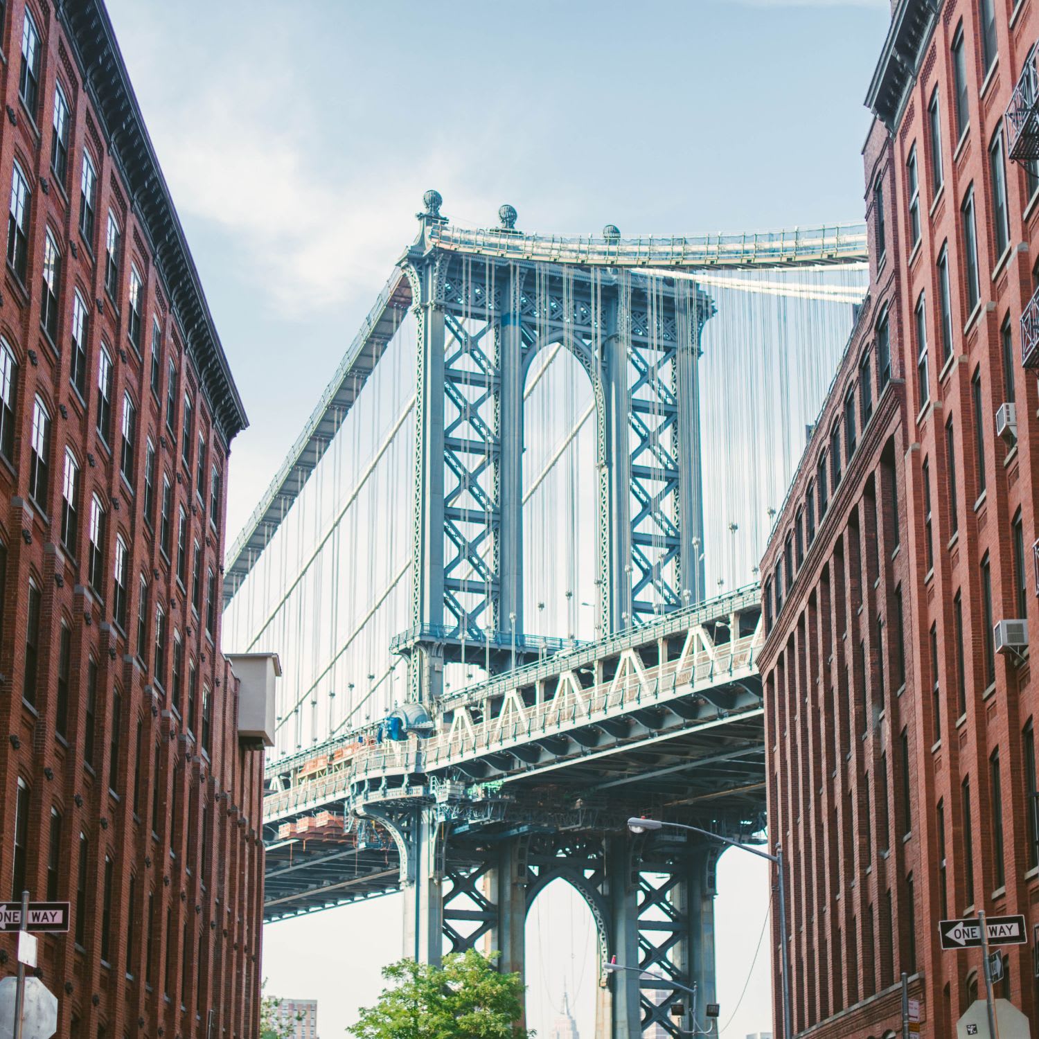 Manhattan Bridge framed by red-brick buildings, viewed from below on a bright day.