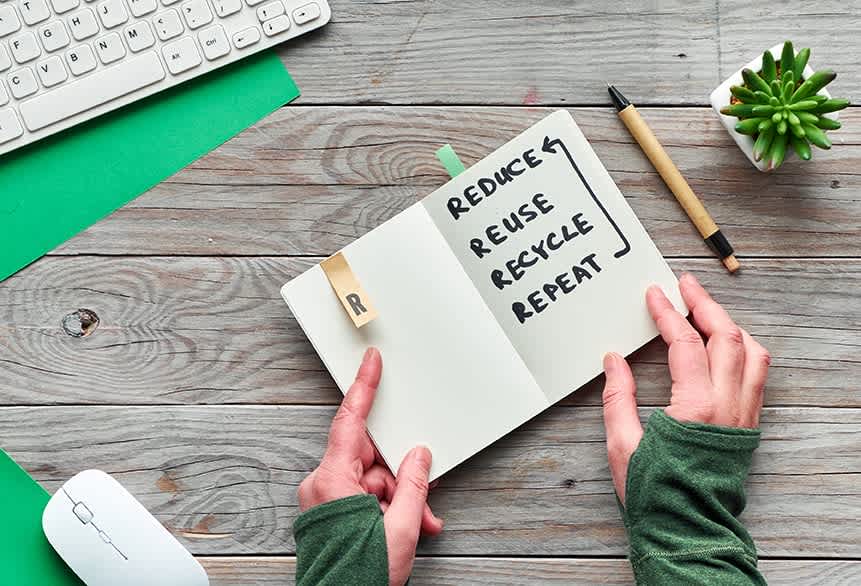 Concept zero waste conscious consumption flat lay, Top view of hands holding trendy moleskin notebook with text "reduce, reuse, recycle, repeat". Wooden table with computer keyboard, mouse and plant
