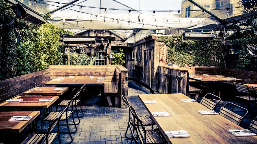 Tipsy Pig’s outdoor seating area bathed in gentle sunlight. Rustic wooden tables and chairs cast intricate shadows on the paved floor, creating a dance of light and dark. The transparent canopy overhead provides shelter while allowing diners to enjoy an open-air experience. Each table is meticulously set, inviting guests to savor a delightful meal amidst lush greenery. 