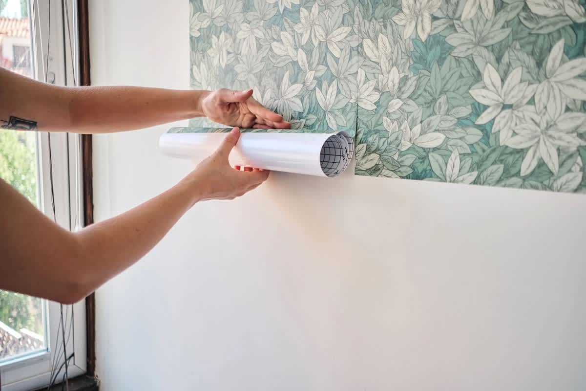 A person is applying floral wallpaper to a wall, holding a roll of wallpaper near the surface.