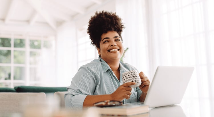 A woman with dark curly hair, smiling as she works on a laptop. She is wearing a light-colored shirt and is seated at a desk in a bright, modern workspace. 