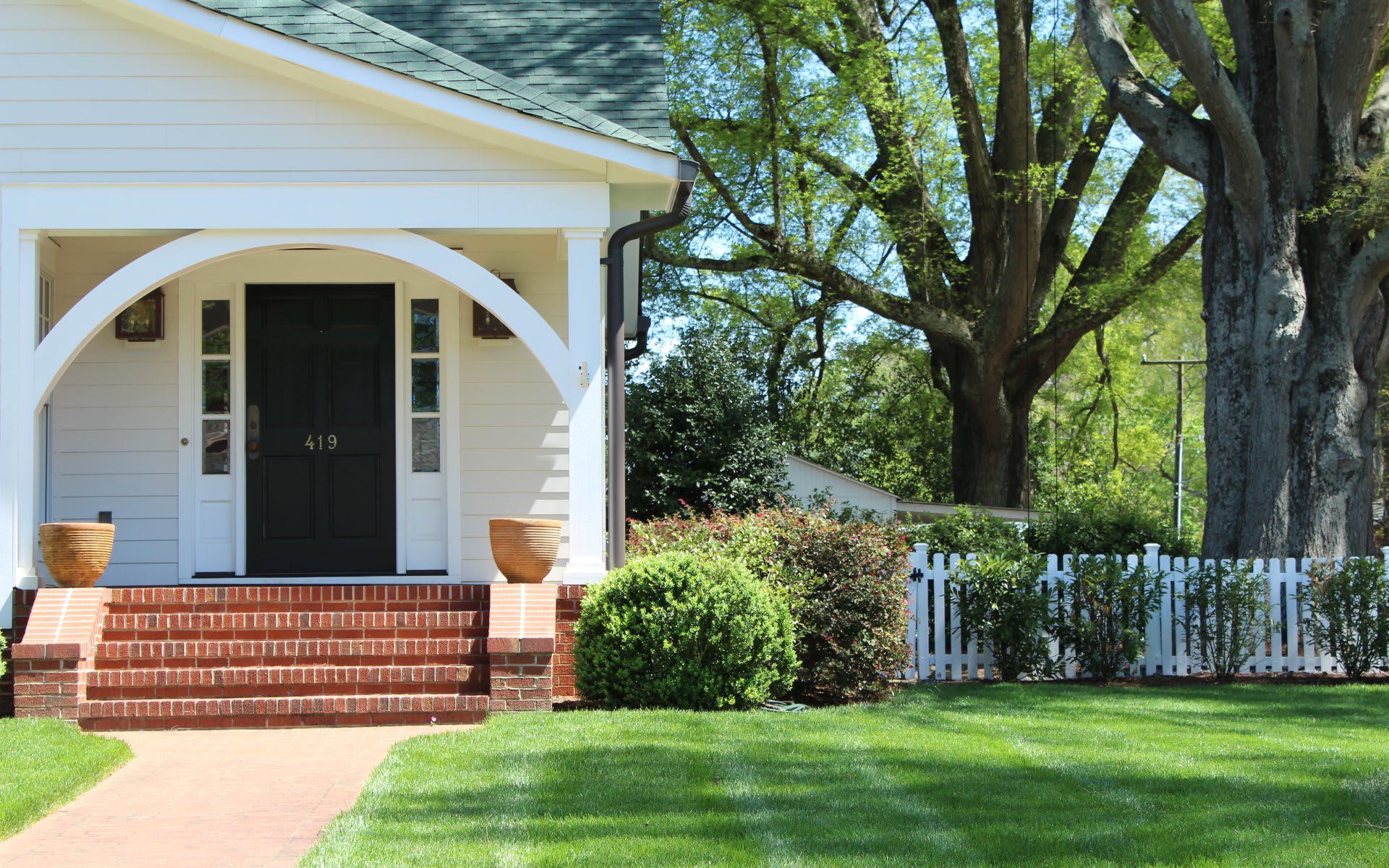 White house with an arched front porch, brick steps, and mature trees beside a picket fence.