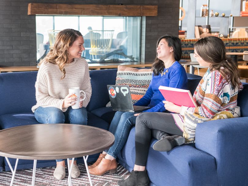 Three women chat on a blue sofa in an office lounge, with a laptop, mug, and pink folder.