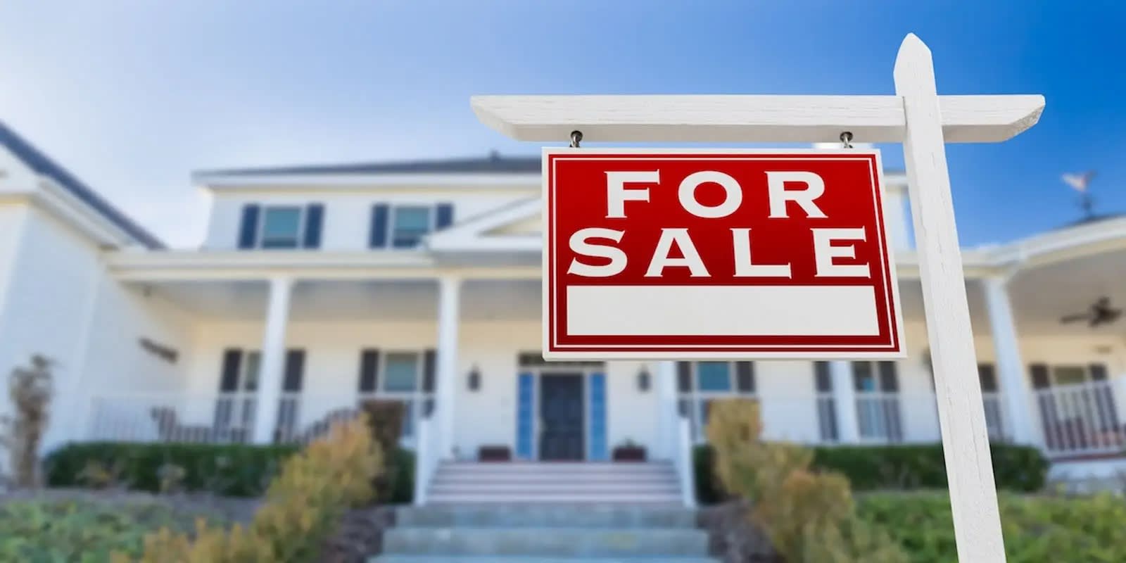 For Sale sign in front of a white house with columns, blurred background.