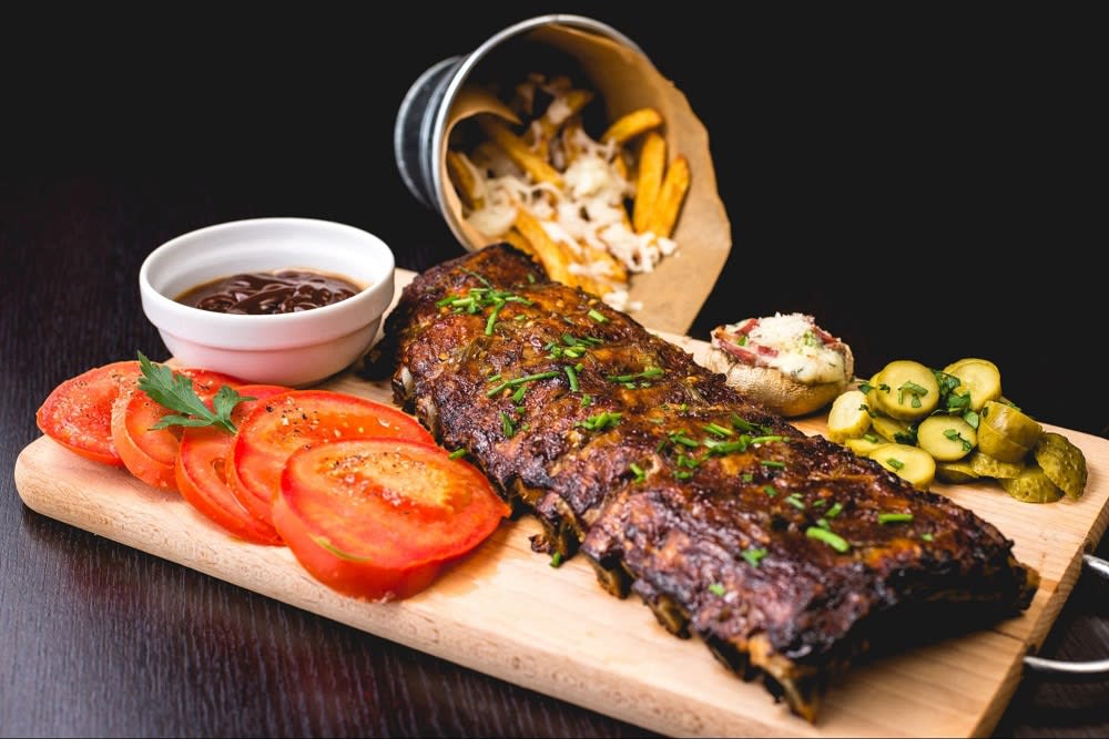 rack of barbequed ribs with sliced tomatoes, pickles, and mushrooms all on a cutting board