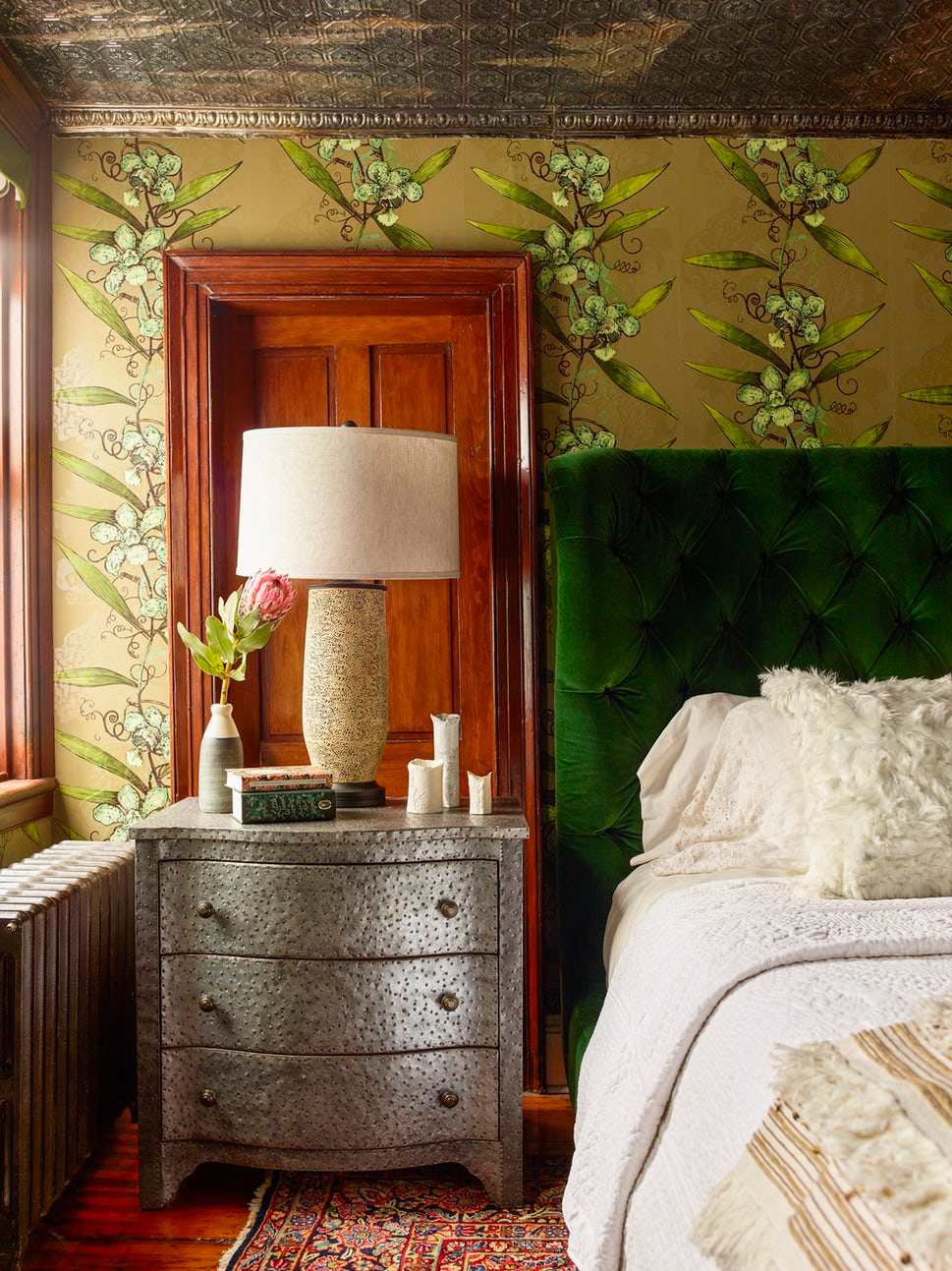 Bedroom with botanical wallpaper, an emerald tufted headboard, and a patterned metal bedside chest.