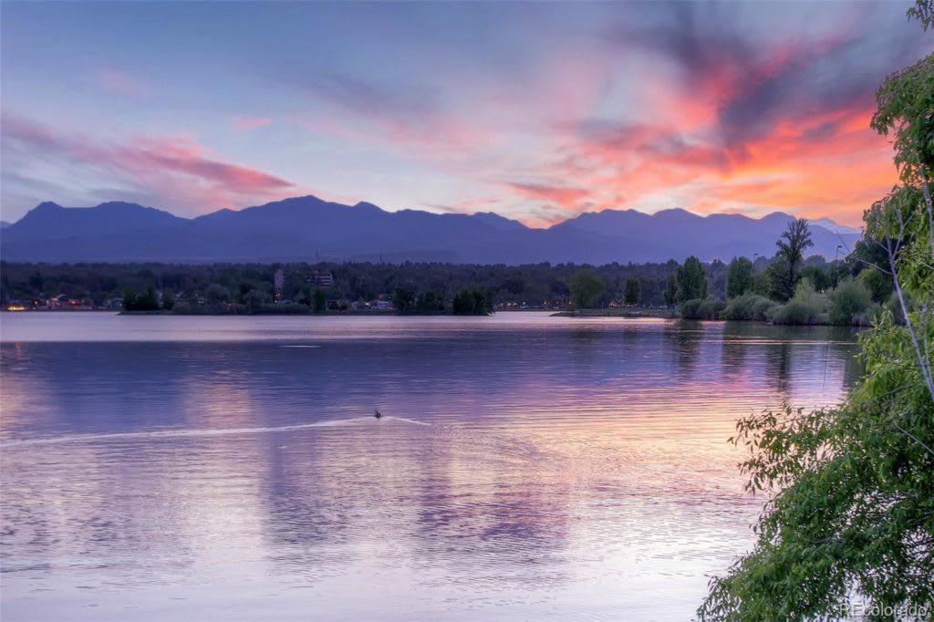 Lake reflecting a pink sunset beneath a mountain range, with trees along the shore.