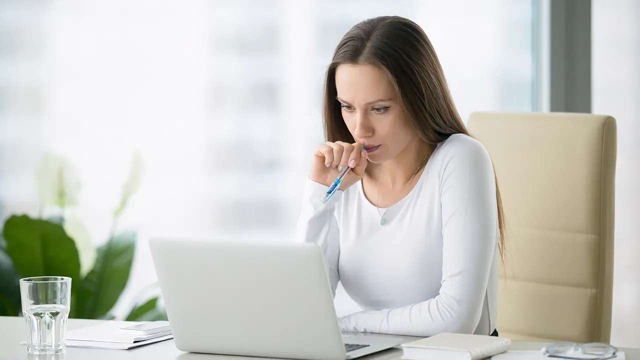 Woman thoughtfully reviewing something on a laptop, holding a pen near her lips.