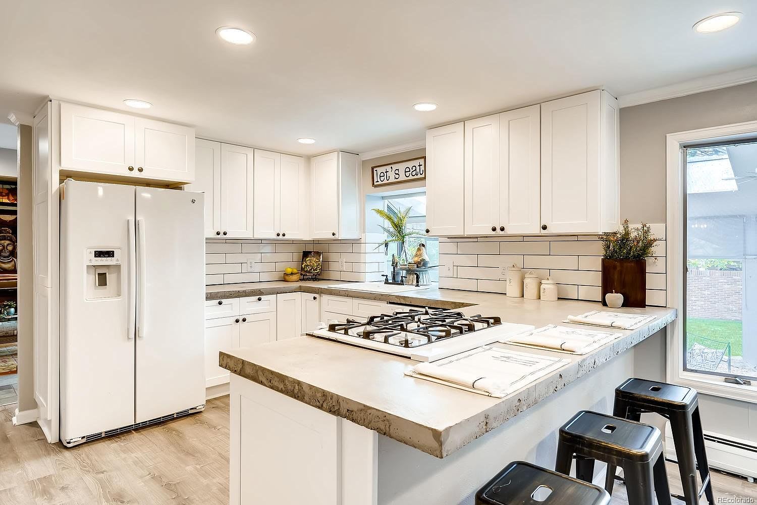Bright white kitchen with a large stone island, gas cooktop, subway tile backsplash, and black stools.