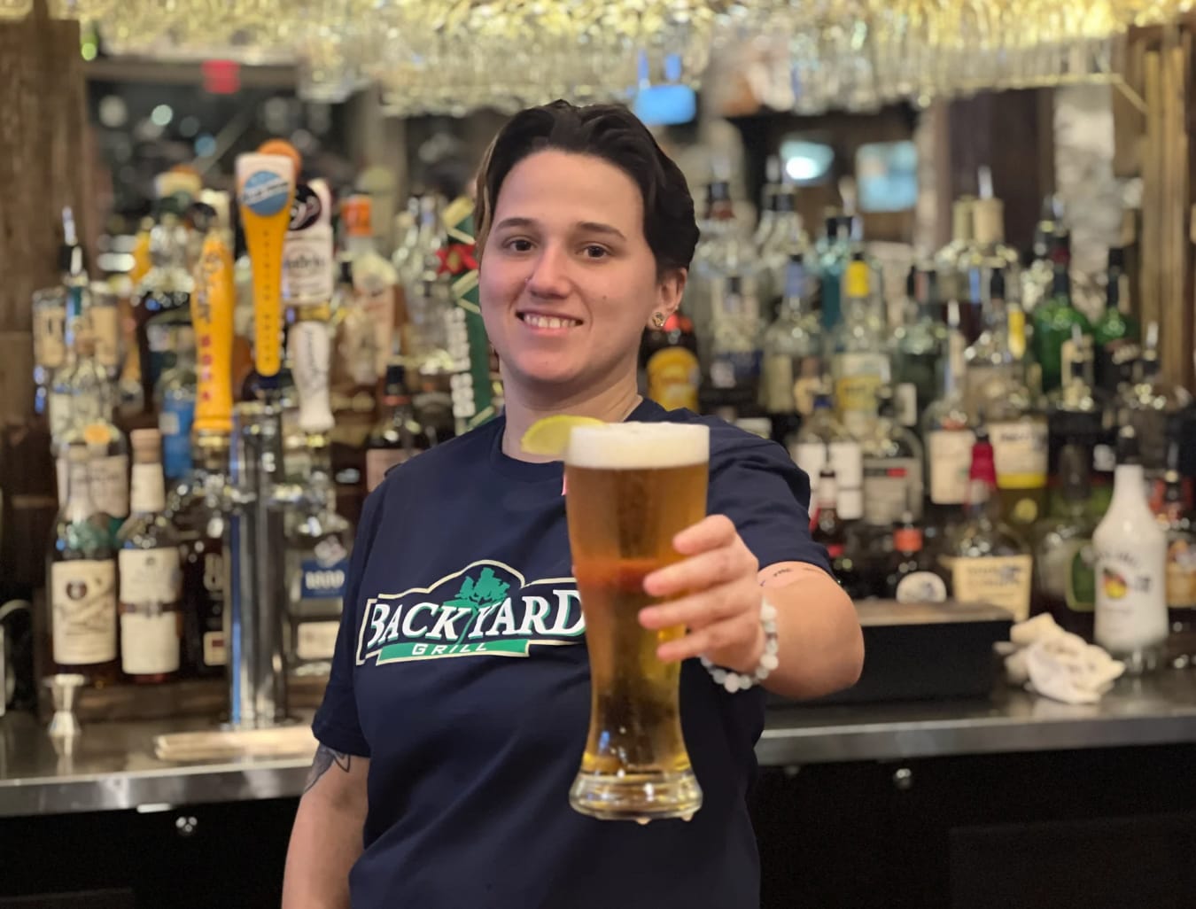 Smiling person offers a pint of amber beer at a bar lined with bottles and taps.