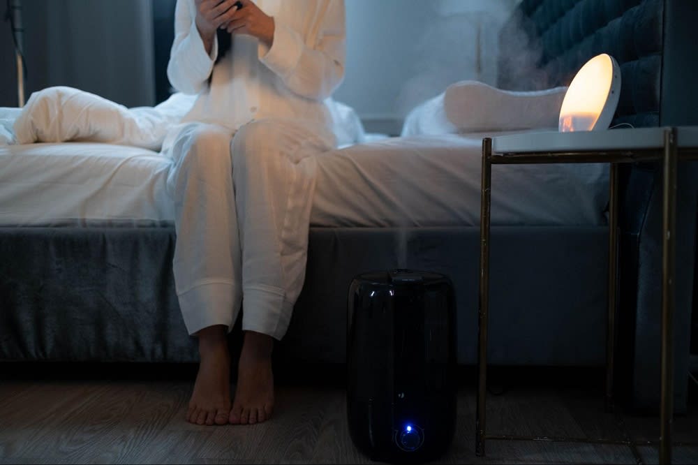 A person in white pajamas sits on a bed, using a phone. A humidifier emits steam beside a small lamp on a nightstand.