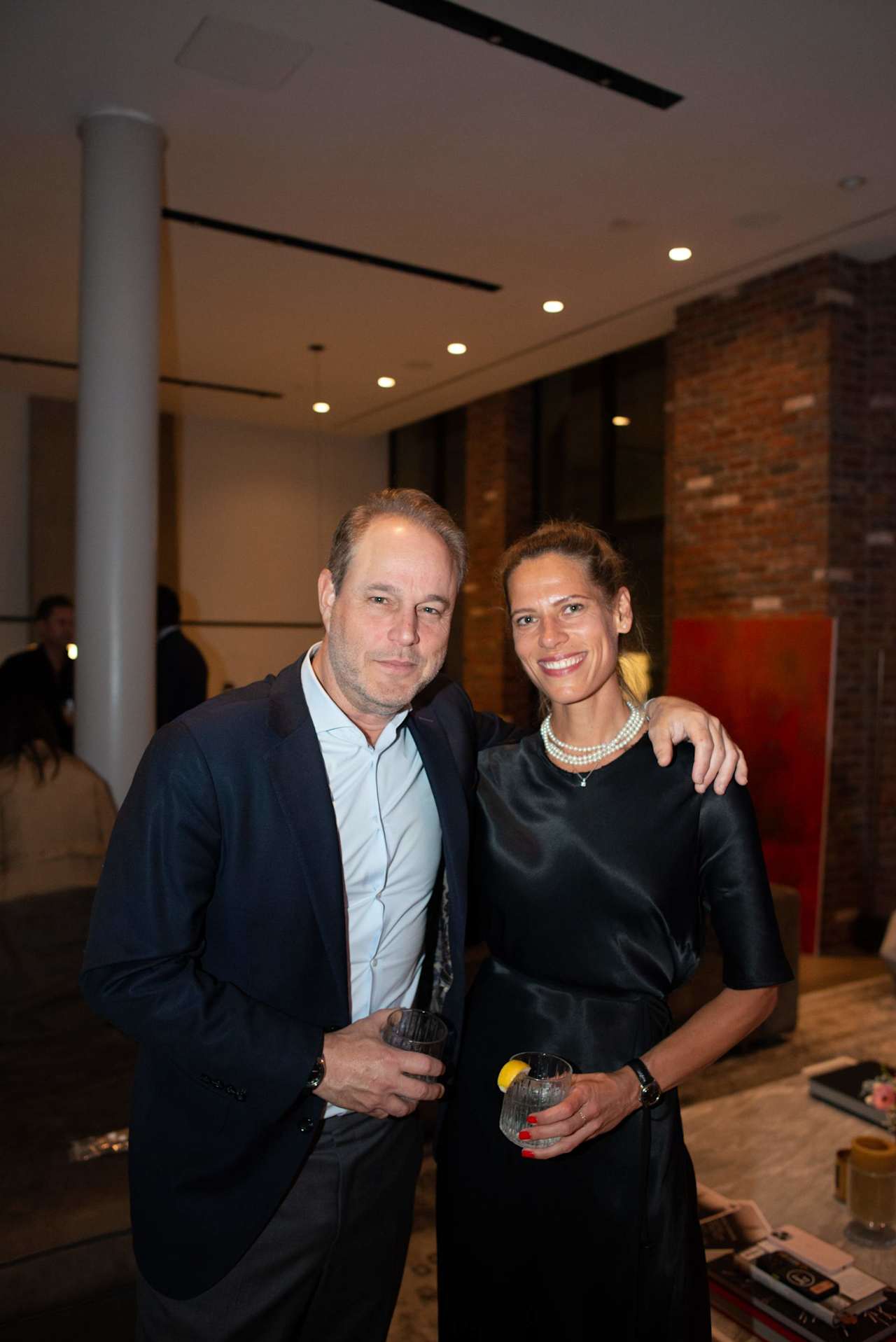 A man in a dark suit and a woman in a black dress pose together holding drinks at an indoor event.