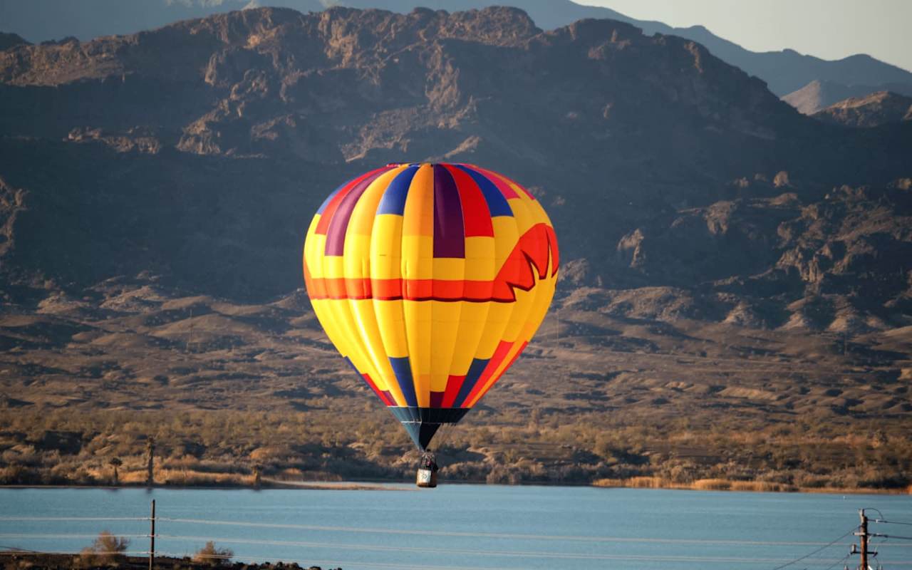 Colorful hot air balloon drifts serenely over calm lake with distant mountains.