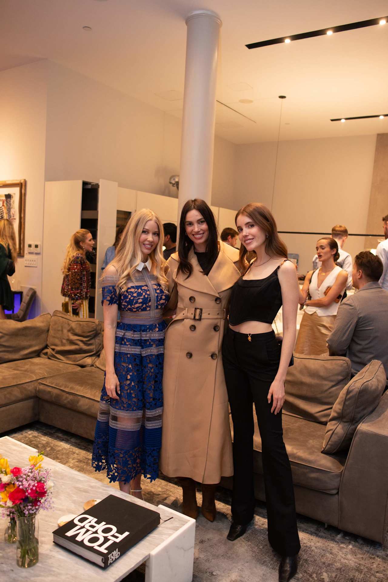 Three women pose at an event in a modern lounge, with guests and sofas behind them.