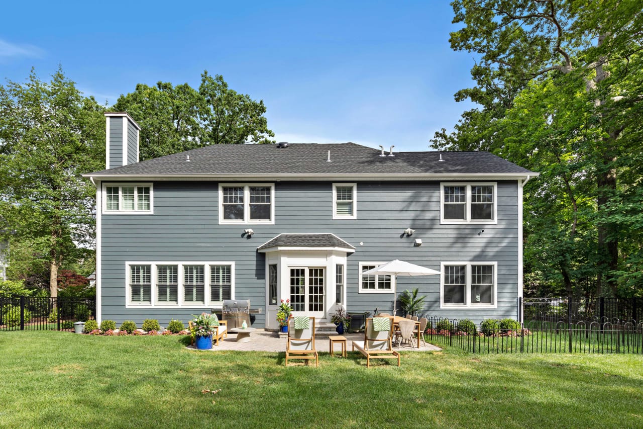 Backyard view of a gray two-story house with a patio area, outdoor seating, and surrounding trees and greenery.