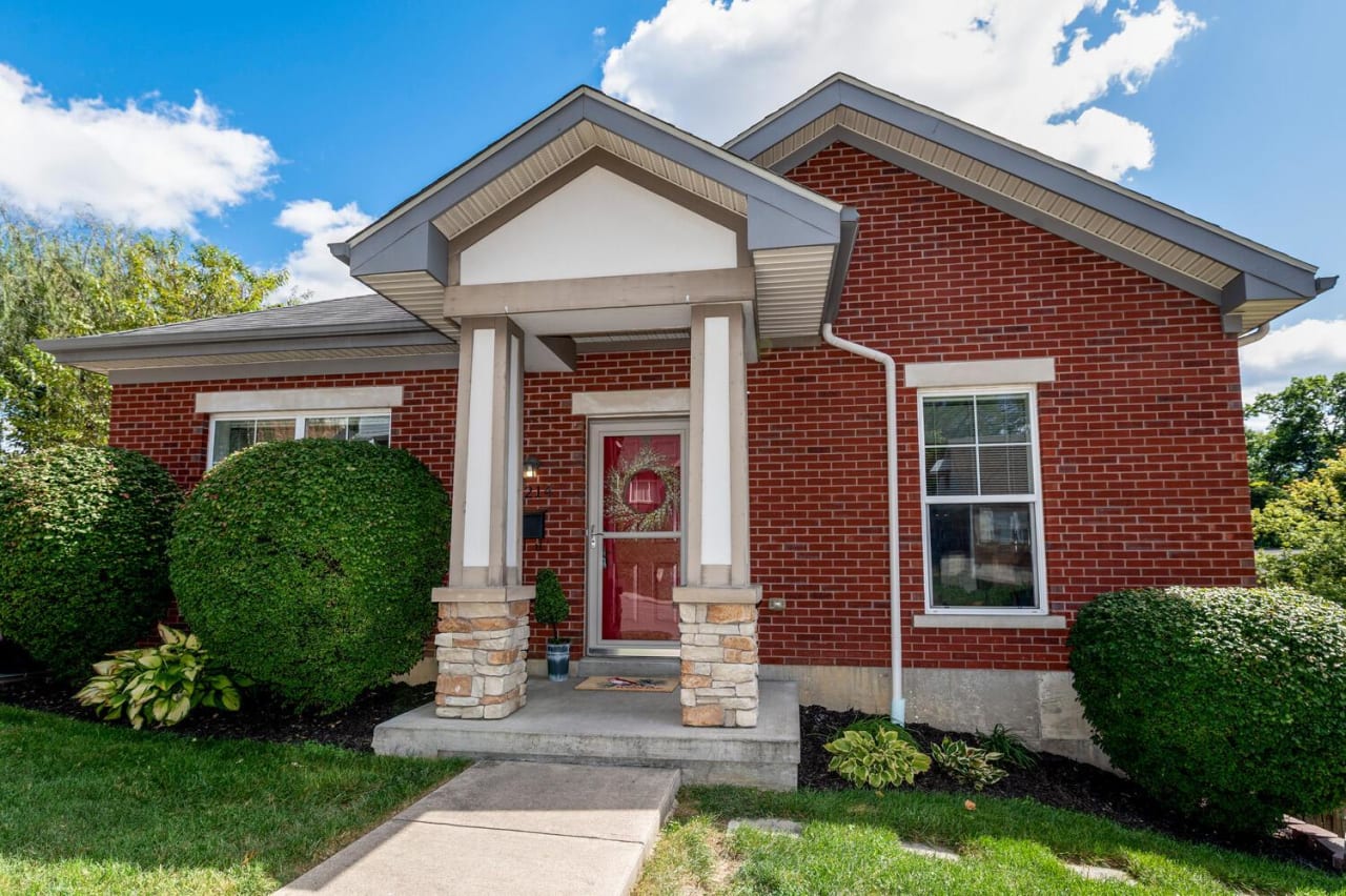 A red brick house with a red door and a sidewalk in front of it. The house is surrounded by bushes and trees.